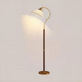 A Clérachic floor lamp with a white pleated shade, curved brass arm, walnut stem and brass base, lit against a pale wall.