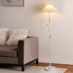 Clérachic floor lamp in white with a pleated shade, lit beside a taupe sofa with fluffy cushions on a woven rug.
