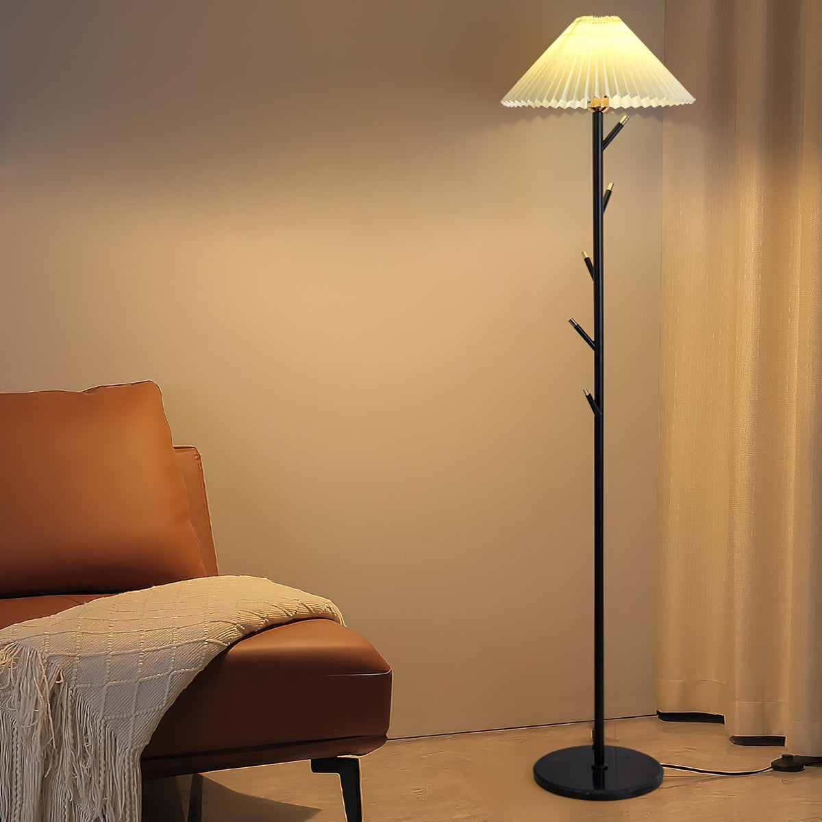 Black Clérachic floor lamp with a pleated shade and short branch hooks, glowing beside a tan leather chair and cream curtain.