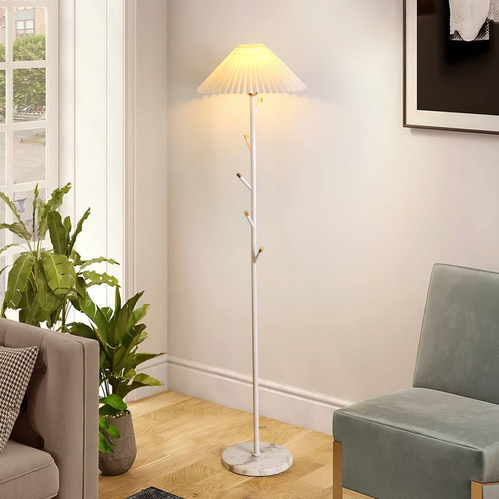 A white Clérachic floor lamp stands by a window in a bright living room, next to a sage-green armchair and leafy plants.