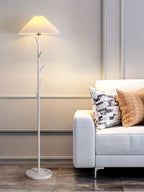 White Clérachic floor lamp with a pleated shade in front of a panelled wall, beside a light grey sofa with patterned cushions.