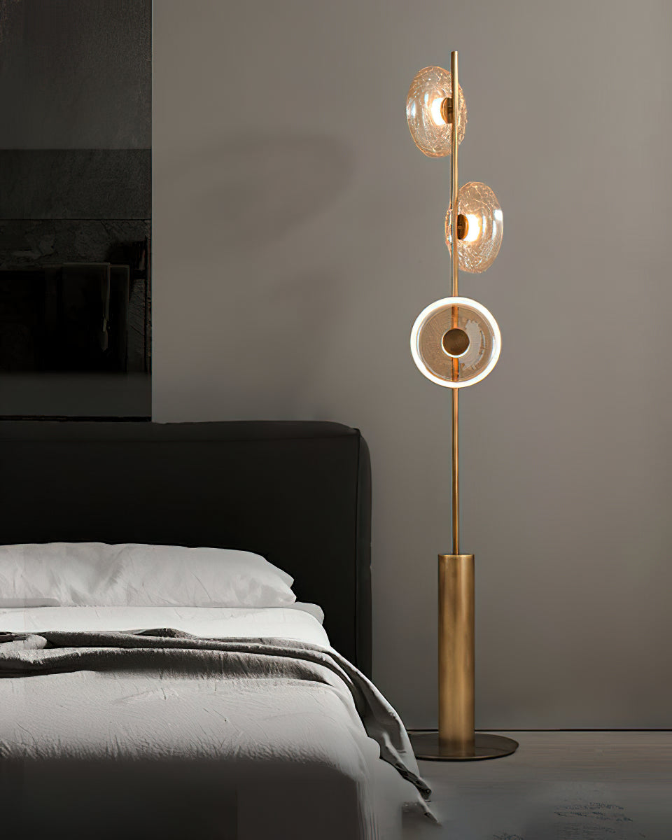 A Clérachic floor lamp with three glass discs stands beside a bed with a dark headboard, the lowest disc lit in a bright ring.