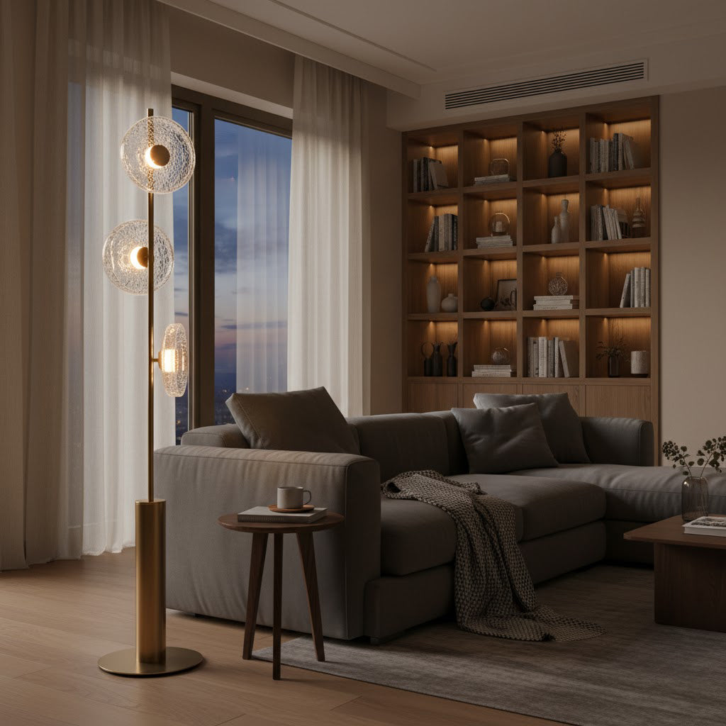 Clérachic floor lamp with three glass discs beside a grey sectional sofa, in front of a lit bookshelf and a window at dusk.