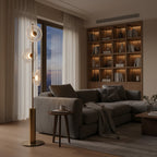 Clérachic floor lamp with three glass discs beside a grey sectional sofa, in front of a lit bookshelf and a window at dusk.