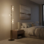 This Clérachic floor lamp stands in a bedroom beside a wooden bedside table, with an abstract landscape print on the wall.
