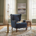 A Clérachic floor lamp with three glass discs beside a navy wingback armchair, in front of sage green walls and tall curtains.
