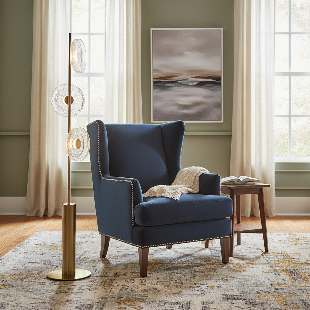 A Clérachic floor lamp with three glass discs beside a navy wingback armchair, in front of sage green walls and tall curtains.