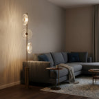 A Clérachic floor lamp beside a grey sofa in a warm room, its glass discs glowing and casting light patterns on the wall.