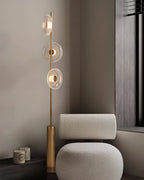 Clérachic floor lamp with its bronze cylinder base beside a cream boucle chair, against a grey wall with a dark window frame.