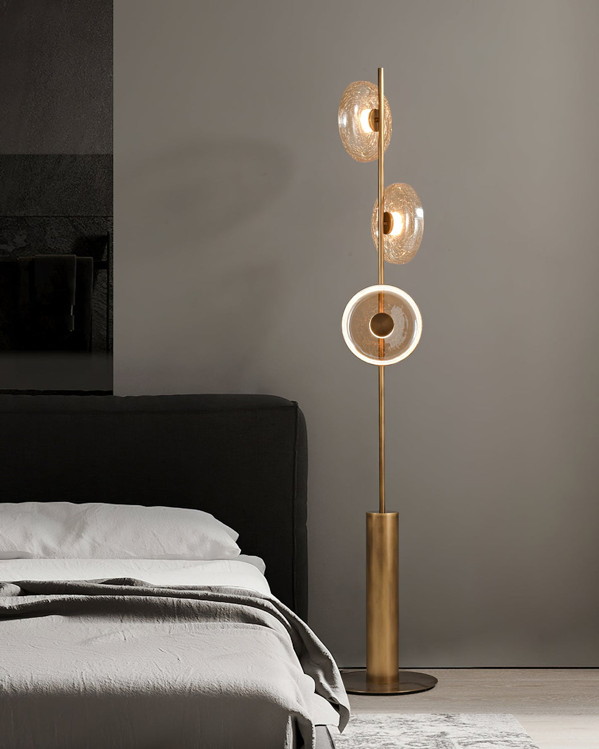 A Clérachic floor lamp beside a bed with a dark headboard and grey bedding, its lowest disc lit as a bright ring.