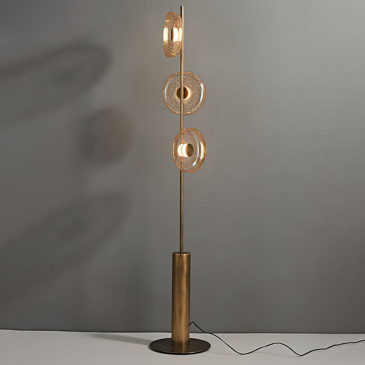 Clérachic floor lamp against a grey wall, its three cracked glass discs warm amber with a cord trailing across the floor.