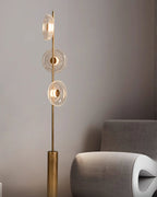 A Clérachic floor lamp rises from its bronze cylinder base behind a sculptural grey chair, in a muted room.