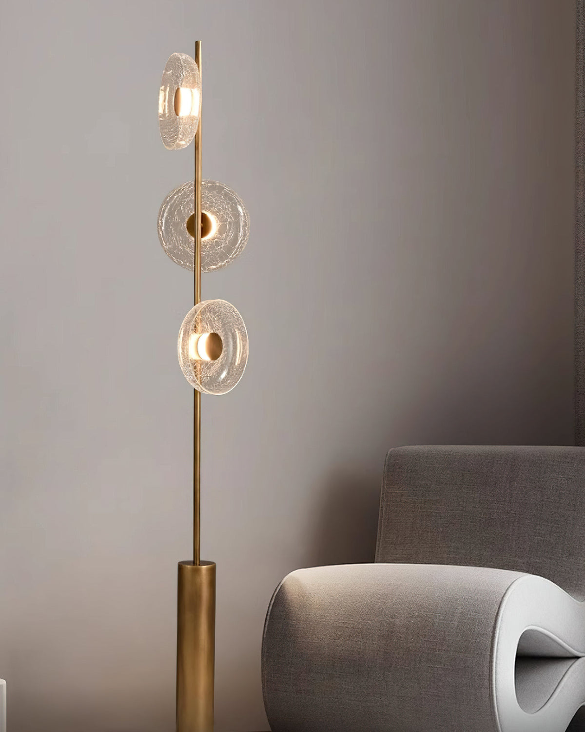 A Clérachic floor lamp rises from its bronze cylinder base behind a sculptural grey chair, in a muted room.