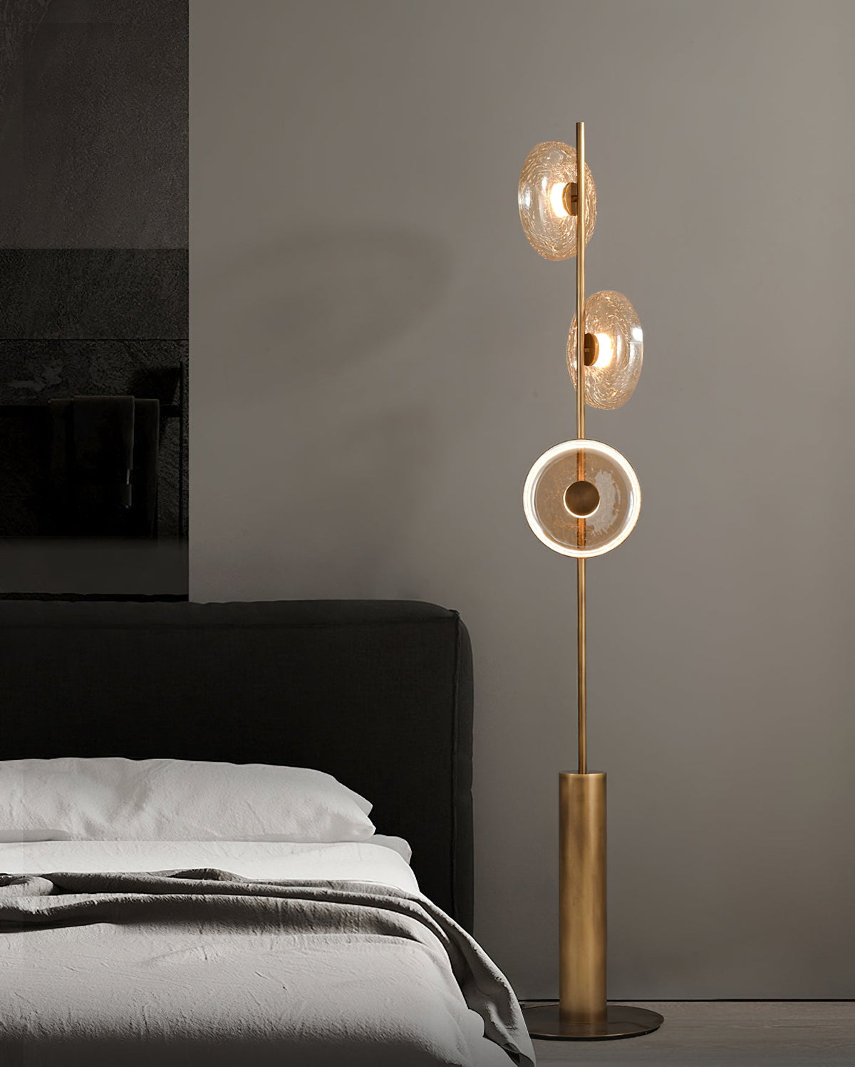 A Clérachic floor lamp with three glass discs stands by a bed with grey bedding, the lowest disc lit as a bright ring.
