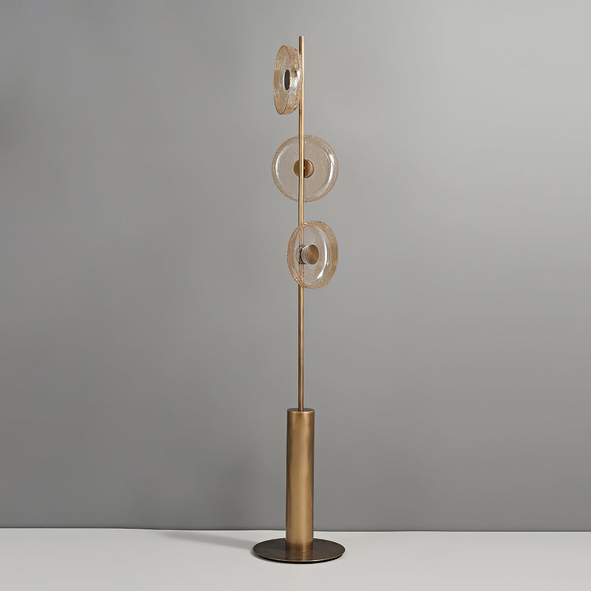 Clérachic floor lamp, unlit, against a grey wall, with three amber-tinted glass discs on a slim stem above a bronze base.