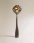 Clérachic floor lamp with a dark gunmetal cone stem and a rippled grey glass-look shade lit warm on a plain backdrop.