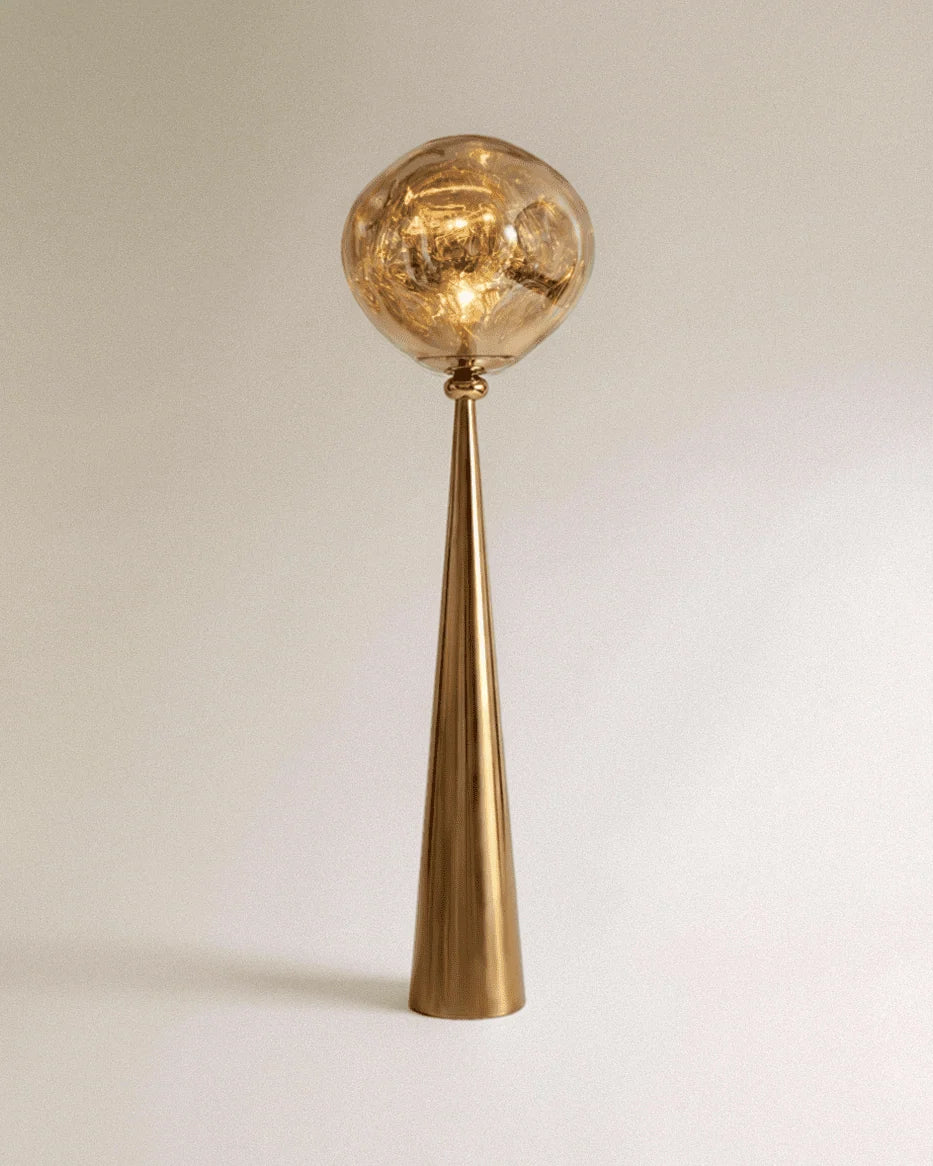 Clérachic floor lamp with a polished gold cone stem and a rippled amber-gold shade lit warm on a plain backdrop.