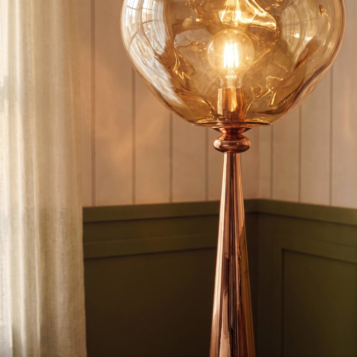 Clérachic rose gold cone stem and amber rippled shade with a visible filament bulb, beside sheer curtains.