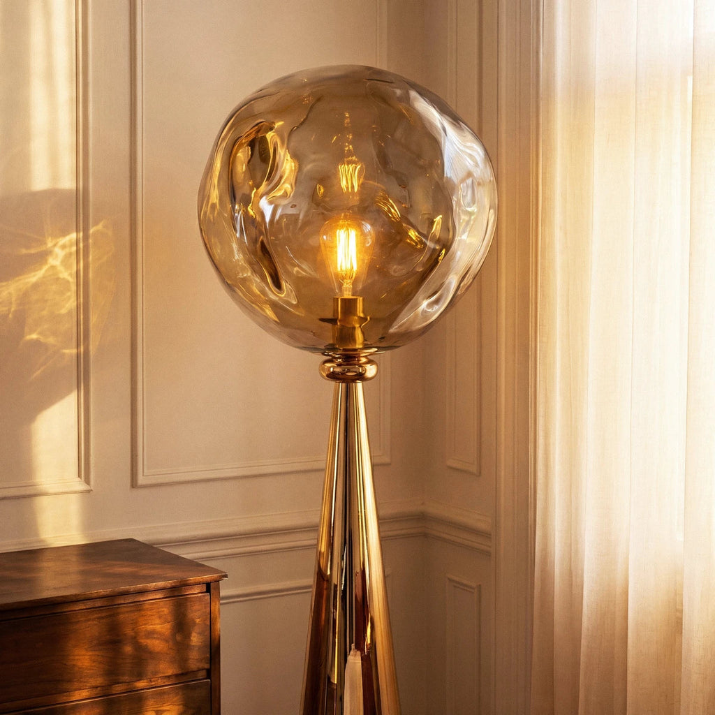 Clérachic gold cone stem and rippled amber shade with a filament bulb glowing, beside a timber chest and sheer curtain.