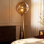 Clérachic gold floor lamp with a rippled amber shade lit warm beside a wooden chest of drawers and a bed.