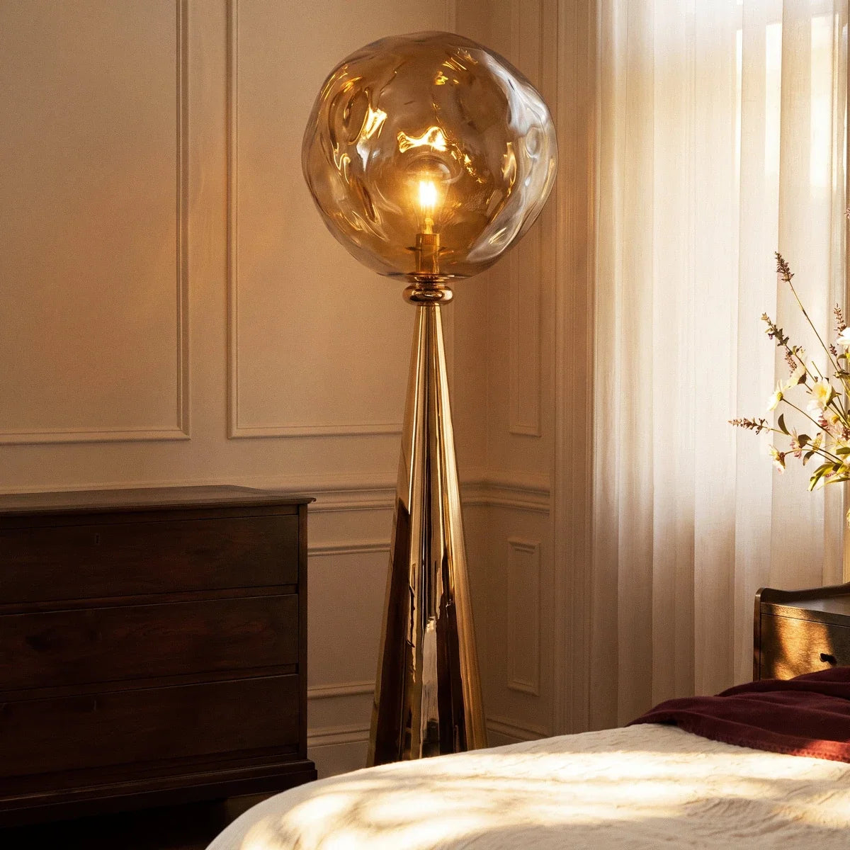 Clérachic gold floor lamp with a rippled amber shade lit warm beside a wooden chest of drawers and a bed.