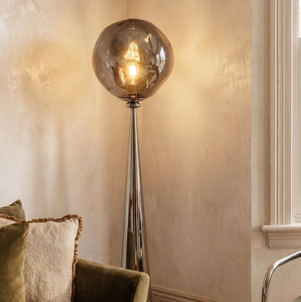 Clérachic floor lamp with a chrome cone stem and rippled grey shade lit warm by a window, beside a green velvet sofa.