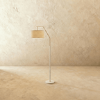 Clérachic slim floor lamp with a matt white stem, round base and brass arched arm carrying a linen drum shade on a limewash wall.