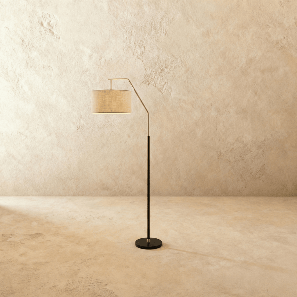 Clérachic floor lamp with a matt black stem and round base, a satin nickel arched arm and a pale linen drum shade, shown in an empty room.