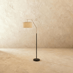 Clérachic floor lamp with a matt black stem and round base, a satin nickel arched arm and a pale linen drum shade, shown in an empty room.