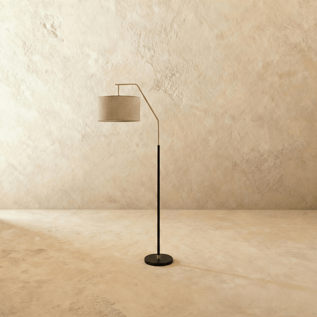 Clérachic arched floor lamp in matt black with a narrow stem, round base and a drum shade in oatmeal fabric against a plaster-textured wall.
