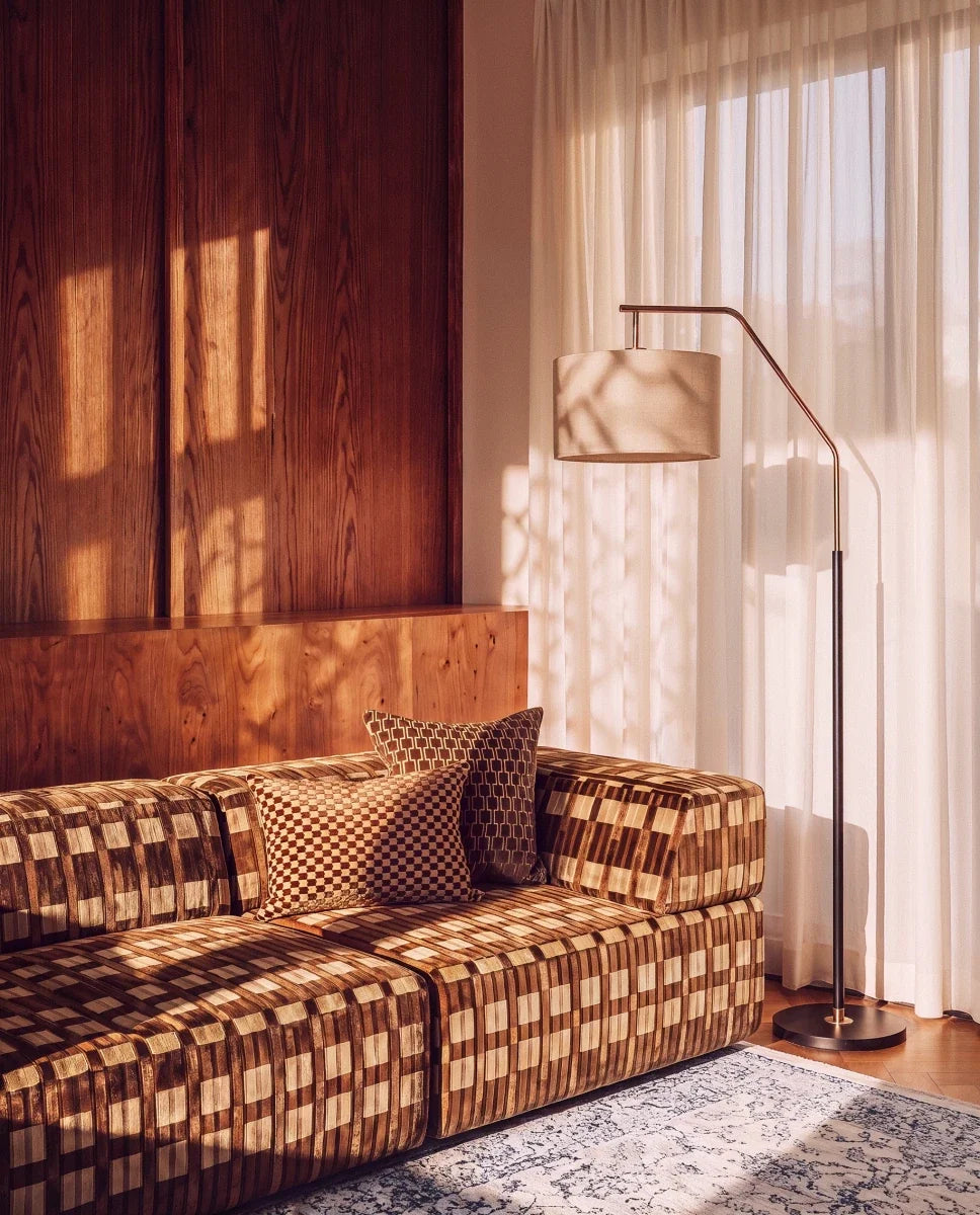Clérachic floor lamp with a dark stem and bronze arm arching over a velvet sofa, its fabric drum shade glowing beside sheer curtains.