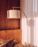 Close view of the Clérachic floor lamp's brass arm and white marble-look stem lifting a linen drum shade above a wood-panelled wall.