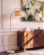 Clérachic white-stemmed floor lamp with a gold arm and drum shade, lighting a panelled wall above a walnut sideboard and books.