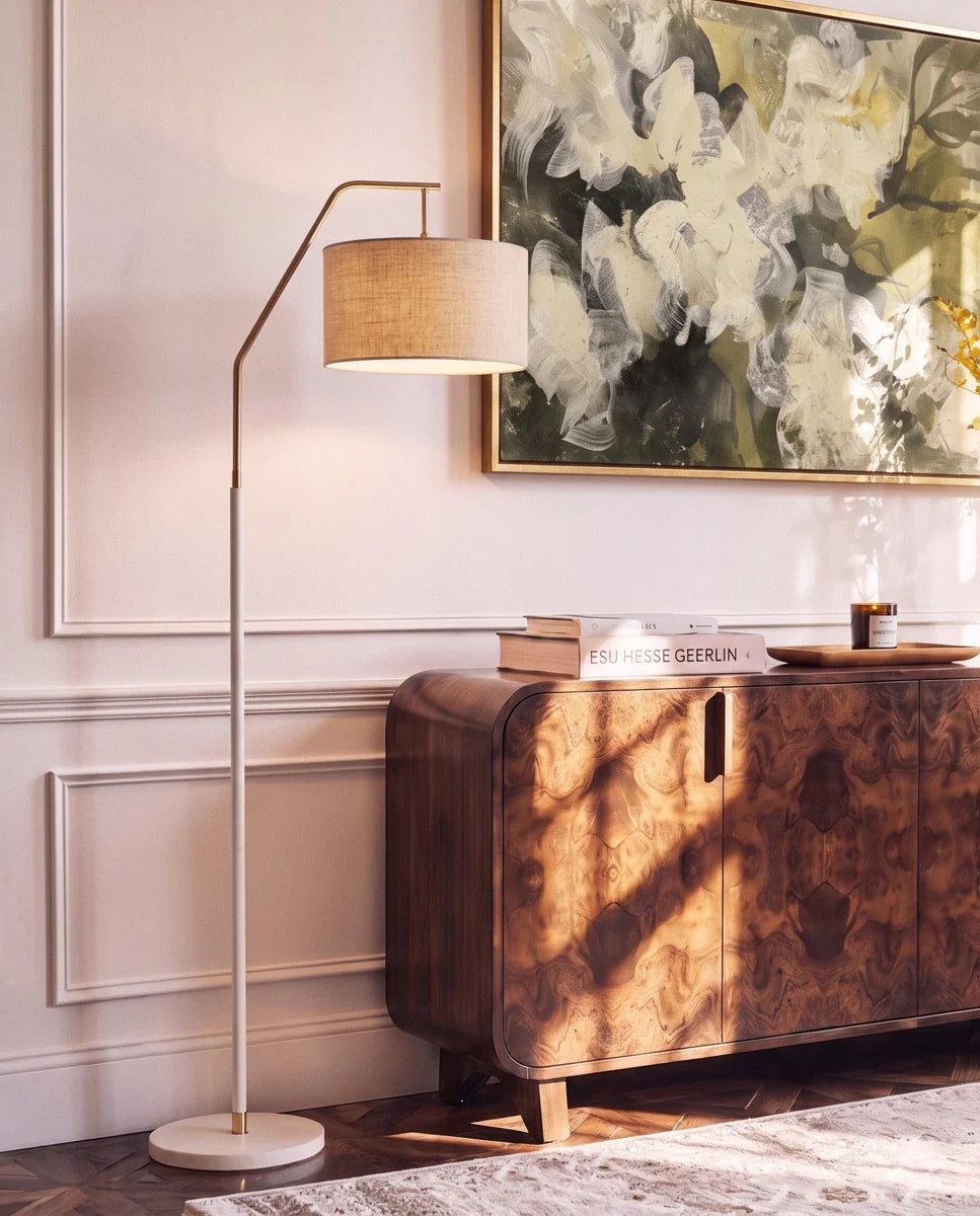 Clérachic white-stemmed floor lamp with a gold arm and drum shade, lighting a panelled wall above a walnut sideboard and books.