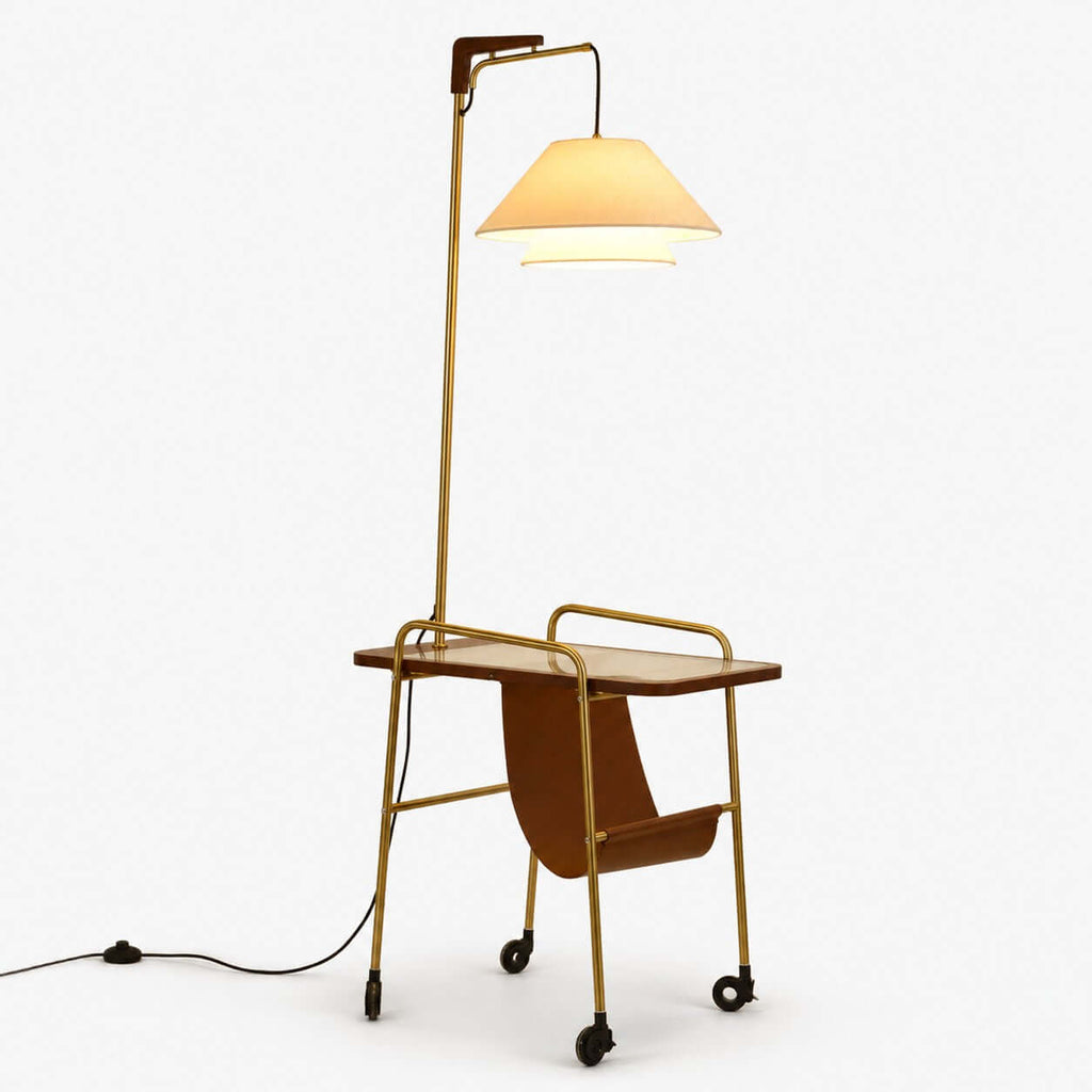 Floor lamp from Clérachic with a brass stem, a tiered cream fabric shade, a wooden tabletop, a leather sling and castors, on white.