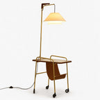 Floor lamp from Clérachic with a brass stem, a tiered cream fabric shade, a wooden tabletop, a leather sling and castors, on white.