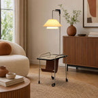 Lit Clérachic floor lamp with a silver frame and a glass tabletop, in front of a fluted sideboard, with a cream bouclé chair at the left.