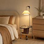 Nickel-framed Clérachic floor lamp beside a bed with cream linen and a pale oak chest of drawers, its shade lit warm.