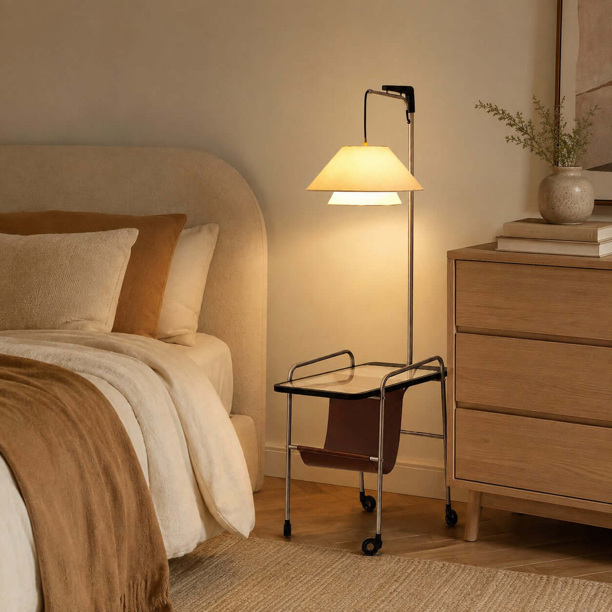 Nickel-framed Clérachic floor lamp beside a bed with cream linen and a pale oak chest of drawers, its shade lit warm.