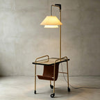 Clérachic floor lamp in brass with a black glass tabletop, a tiered cream shade and a leather sling, against a grey wall.