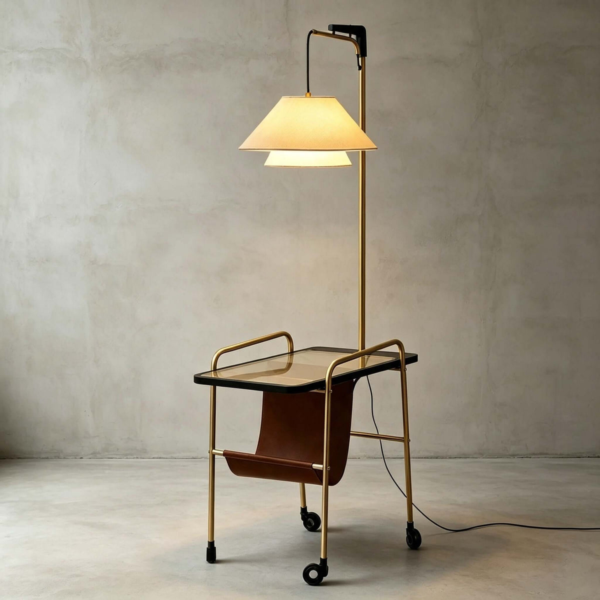 Clérachic floor lamp in brass with a black glass tabletop, a tiered cream shade and a leather sling, against a grey wall.