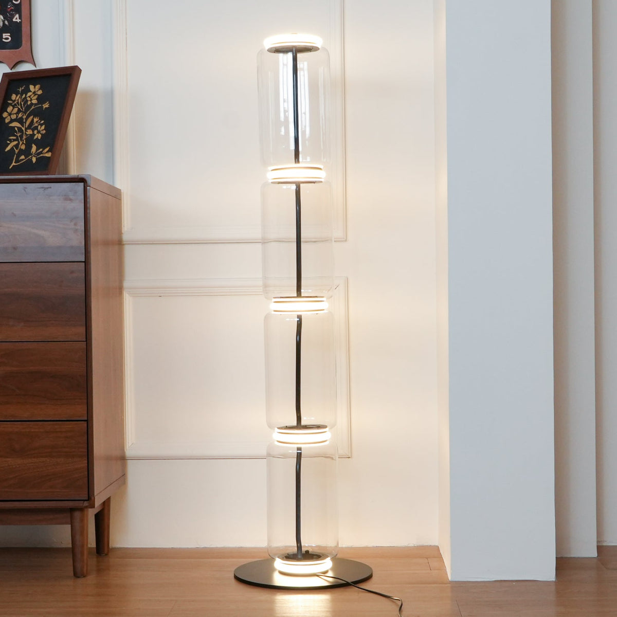 Four-module Clérachic glass floor lamp lit warm beside a walnut chest of drawers, a glowing ring at each joint and at the base.