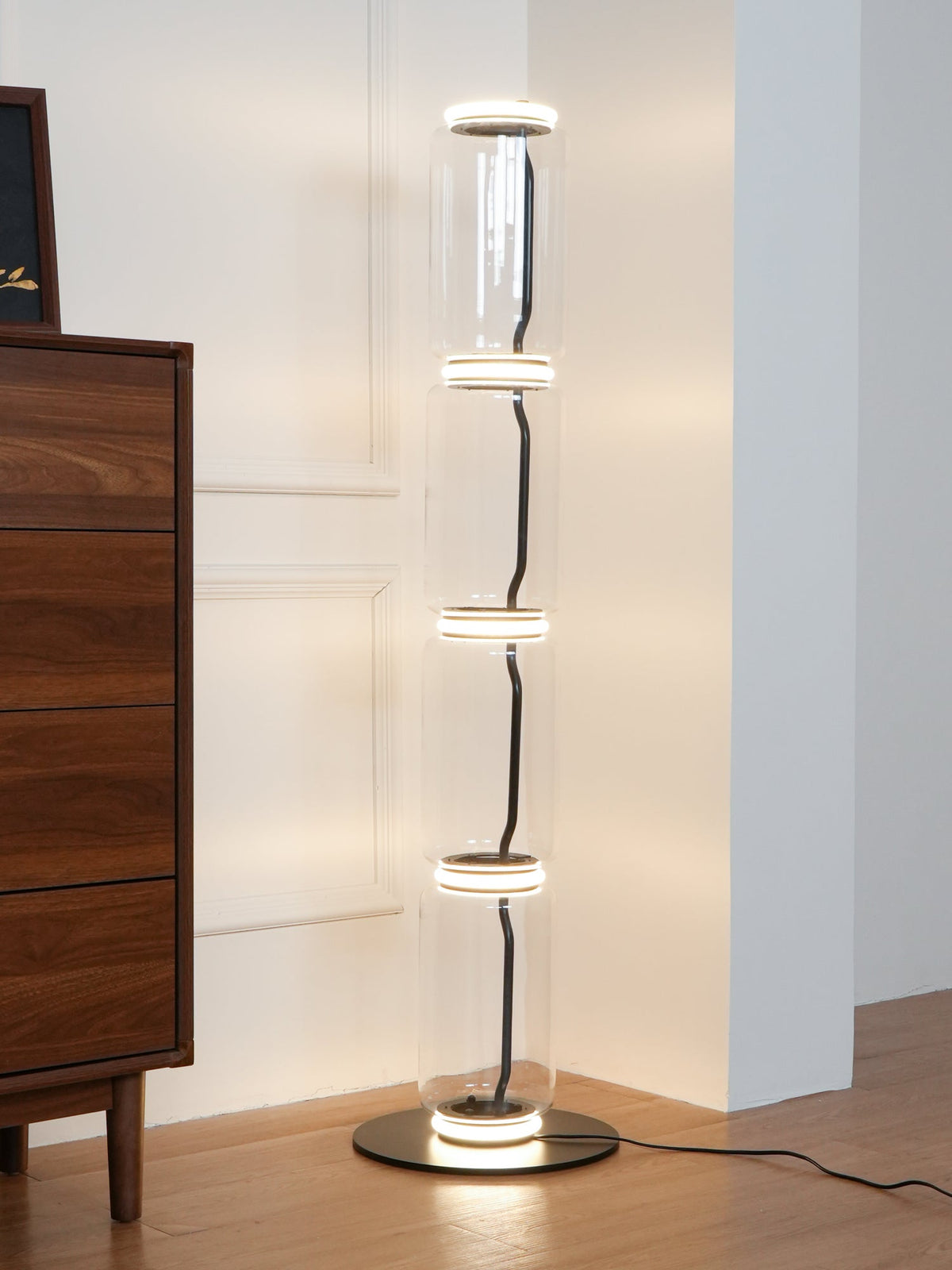 Clérachic glass floor lamp beside a walnut chest in a corner, its stack of clear cylinders glowing at every joint.