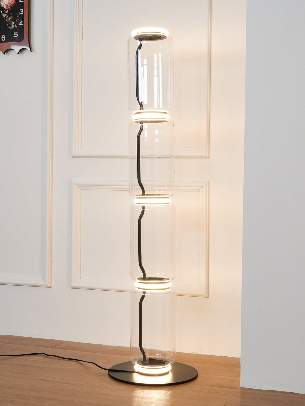 Clérachic glass floor lamp lit warm in front of a white panelled door, with the cord trailing across a wood floor from the black base.