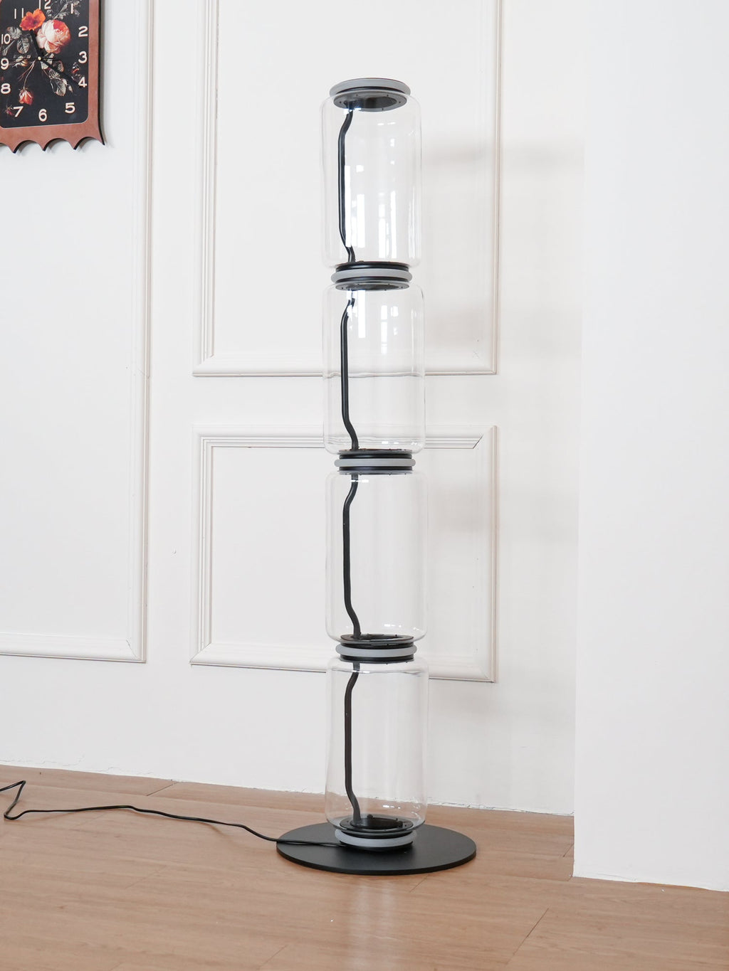 Unlit Clérachic glass floor lamp on a wood floor, tall clear cylinders with black connectors beside a panelled white wall.