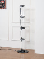 Unlit Clérachic glass floor lamp on a wood floor, tall clear cylinders with black connectors beside a panelled white wall.