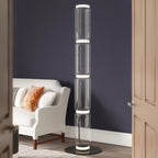 Clérachic glass floor lamp glowing at its joints in a room with deep navy walls, beside a white armchair with orange cushions.