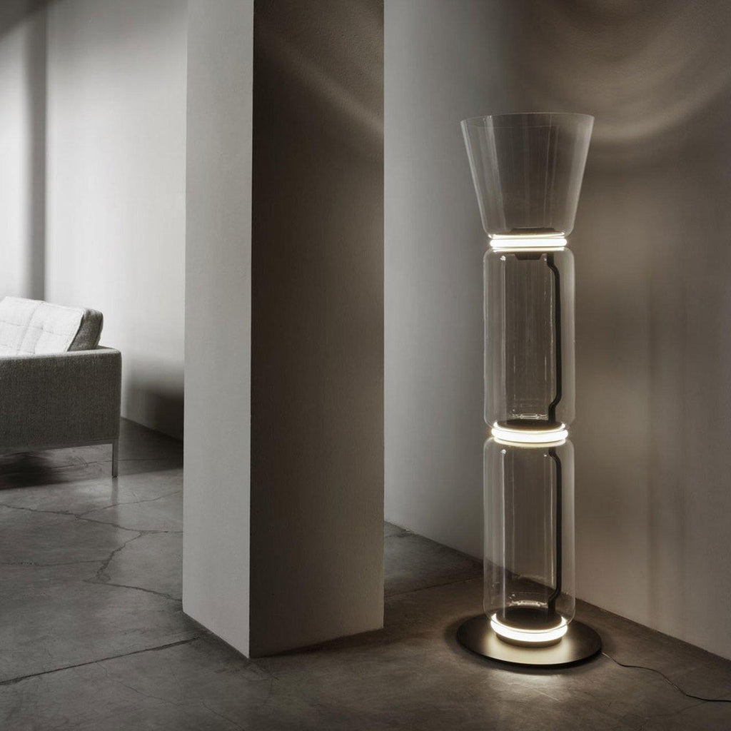 Clérachic glass floor lamp with a cone shade lit at its rings in a dim hall, beside a plain pillar and a grey concrete floor.