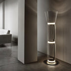 Clérachic glass floor lamp with a cone shade lit at its rings in a dim hall, beside a plain pillar and a grey concrete floor.
