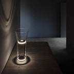 Short Clérachic glass floor lamp with a cone shade on a dark wooden platform, ringed with light against a blue-grey wall.